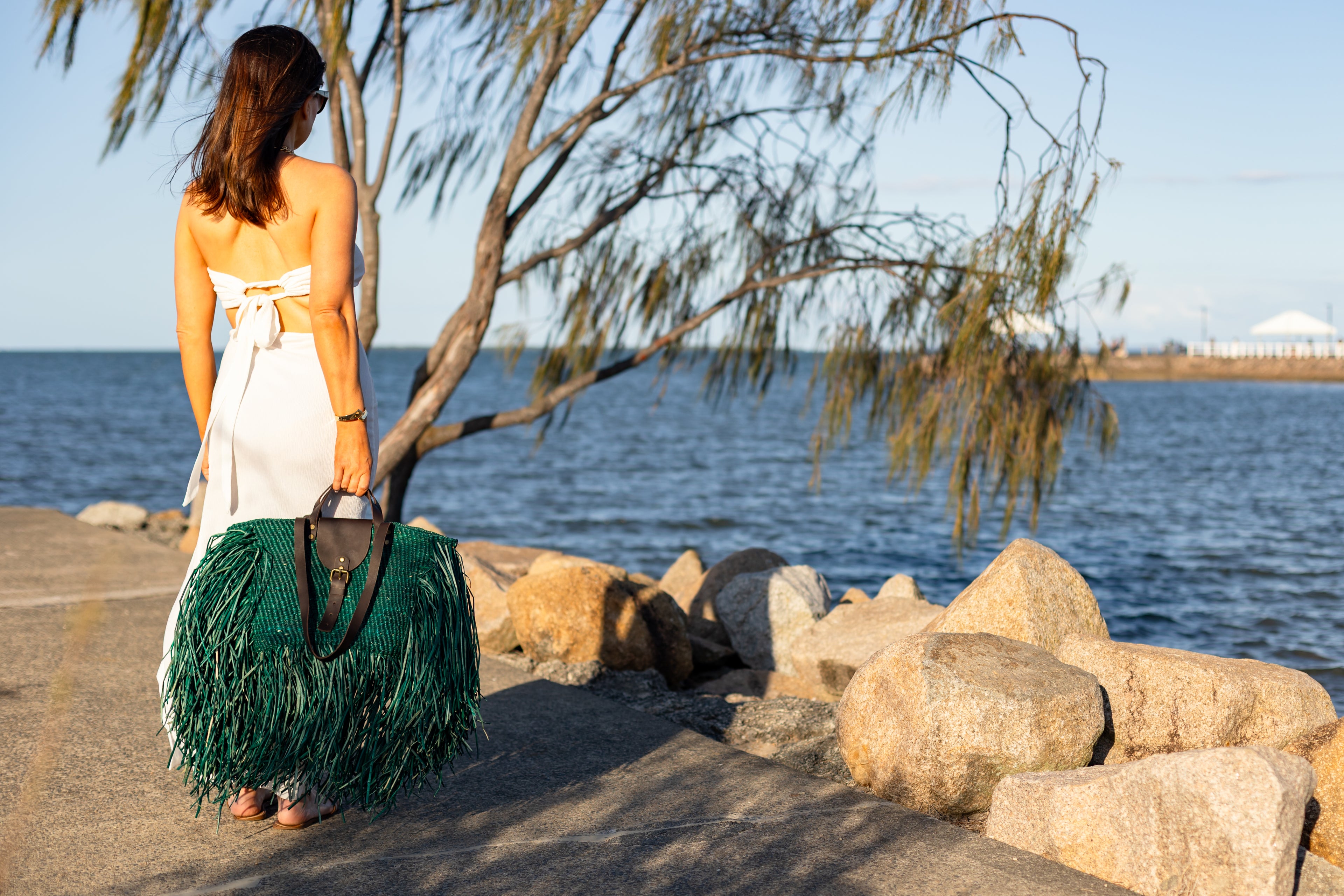 Woman with a green fringed bag standing by a body of water with rocks and trees.