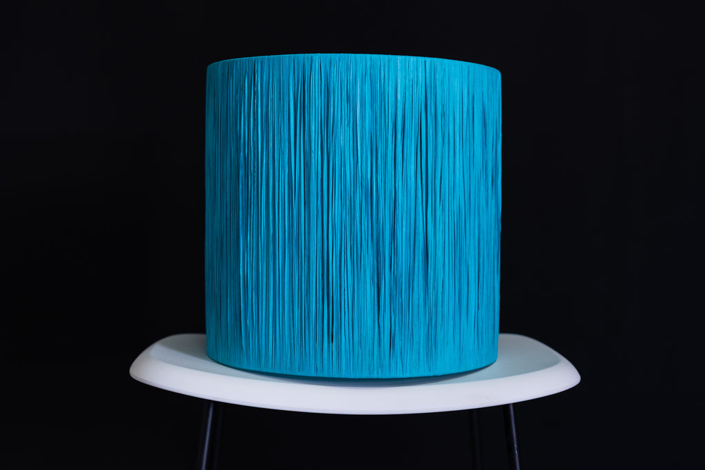 Turquoise raffia wrapped drum lampshade on a white stand against a black background.