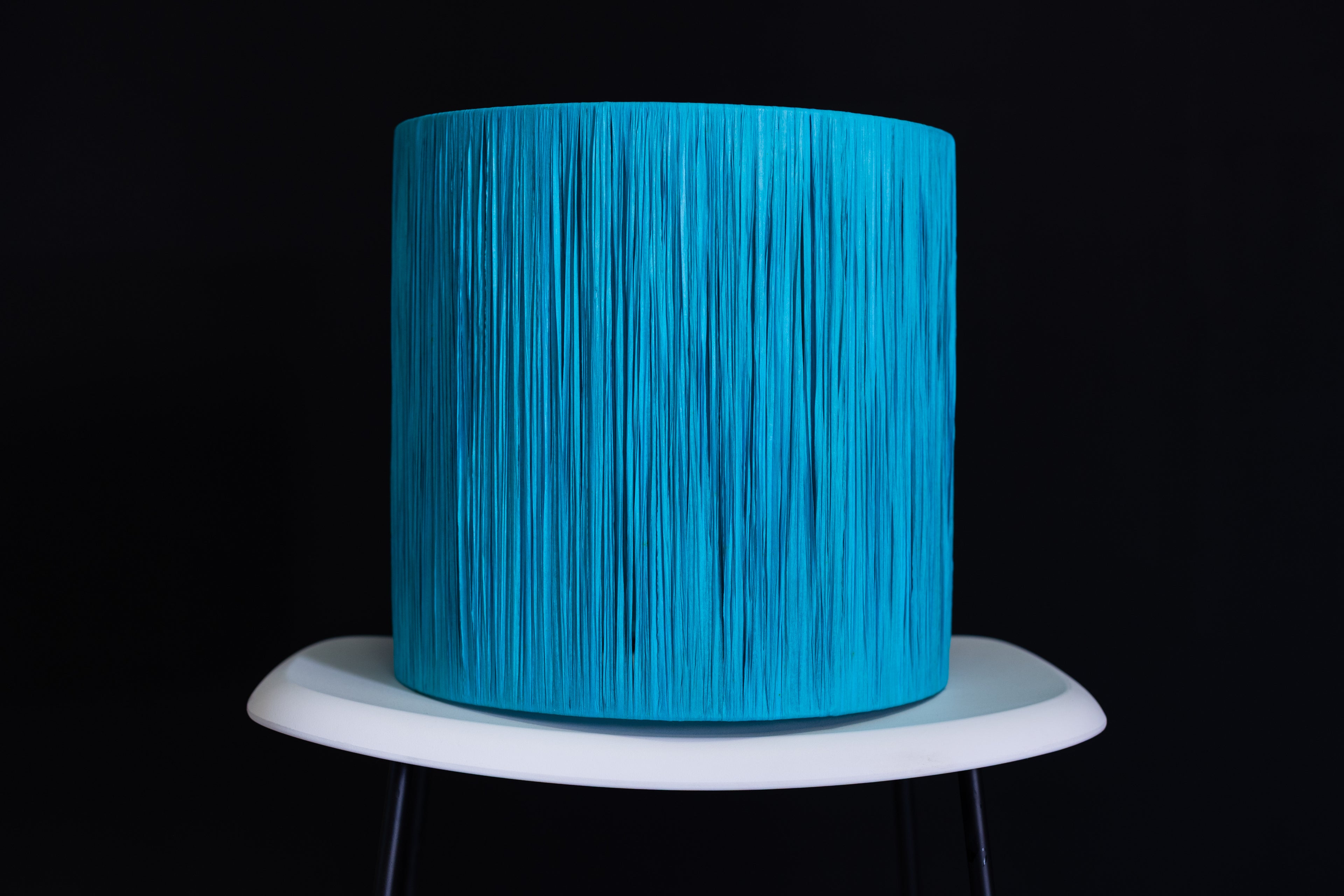 Turquoise raffia wrapped drum lampshade on a white stand against a black background.