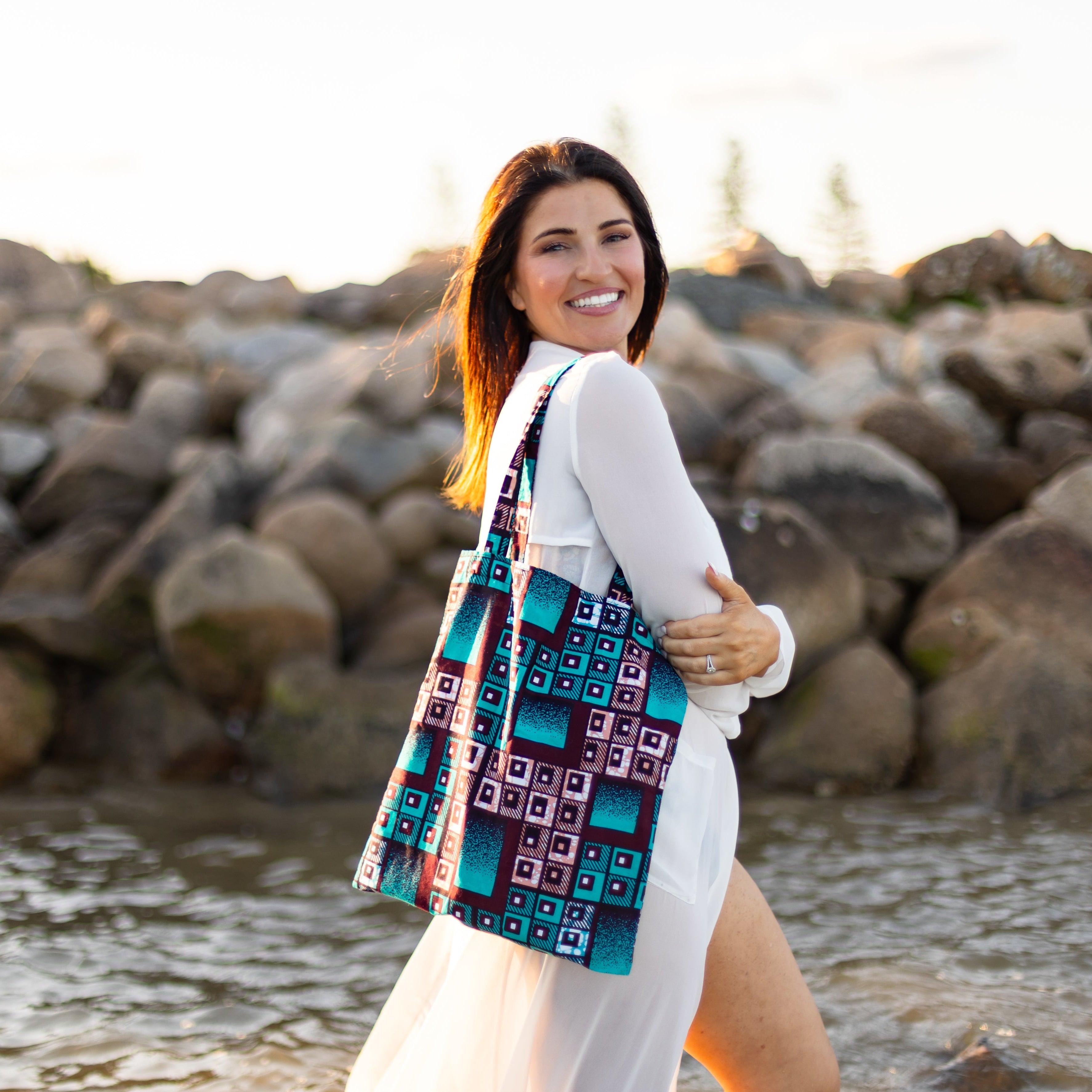 Woman with a patterned bag standing by a body of water with rocks in the background