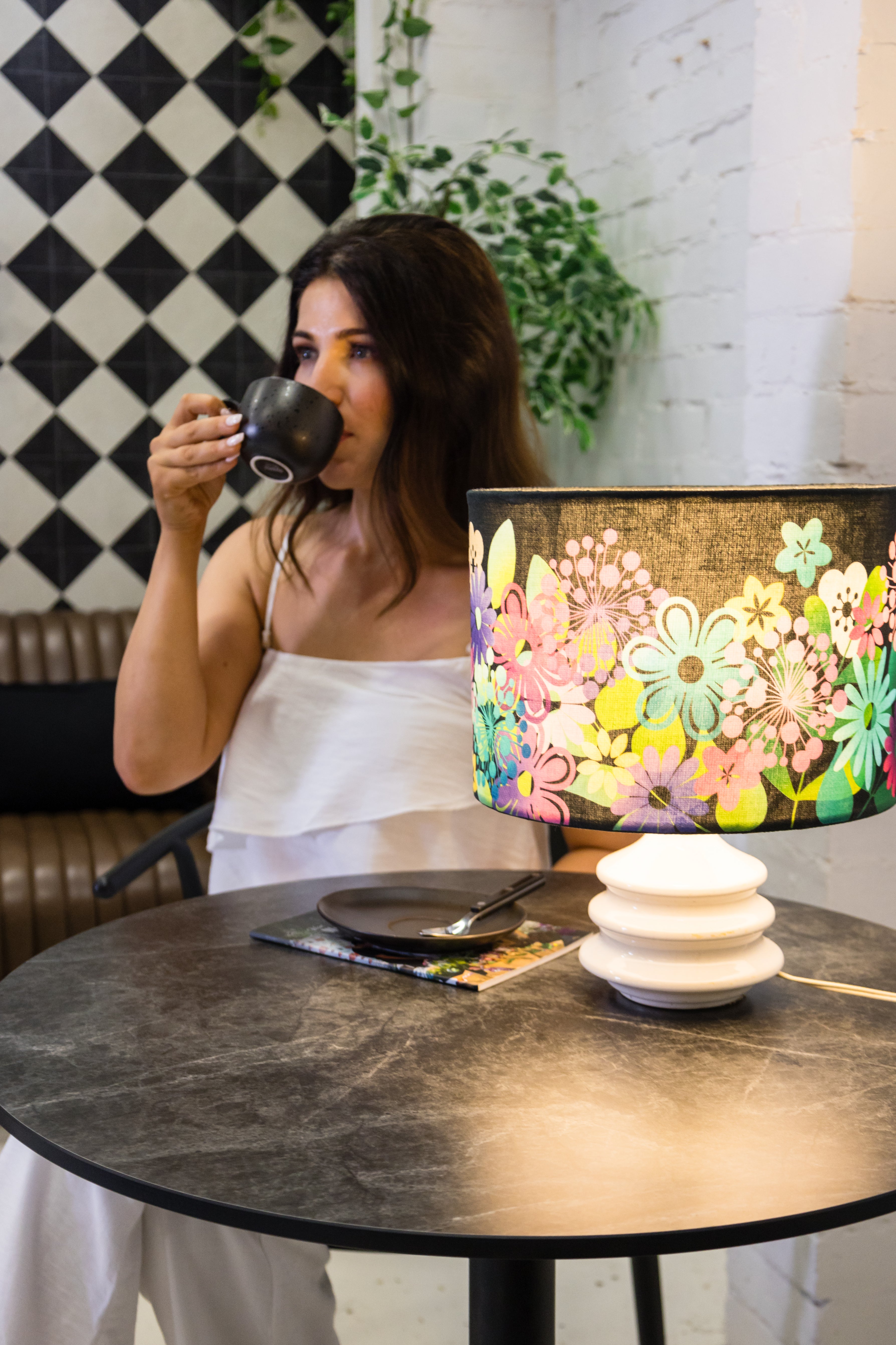 Woman drinking from a coffee cup in a cafe with a colourful lamp and checkered wall.