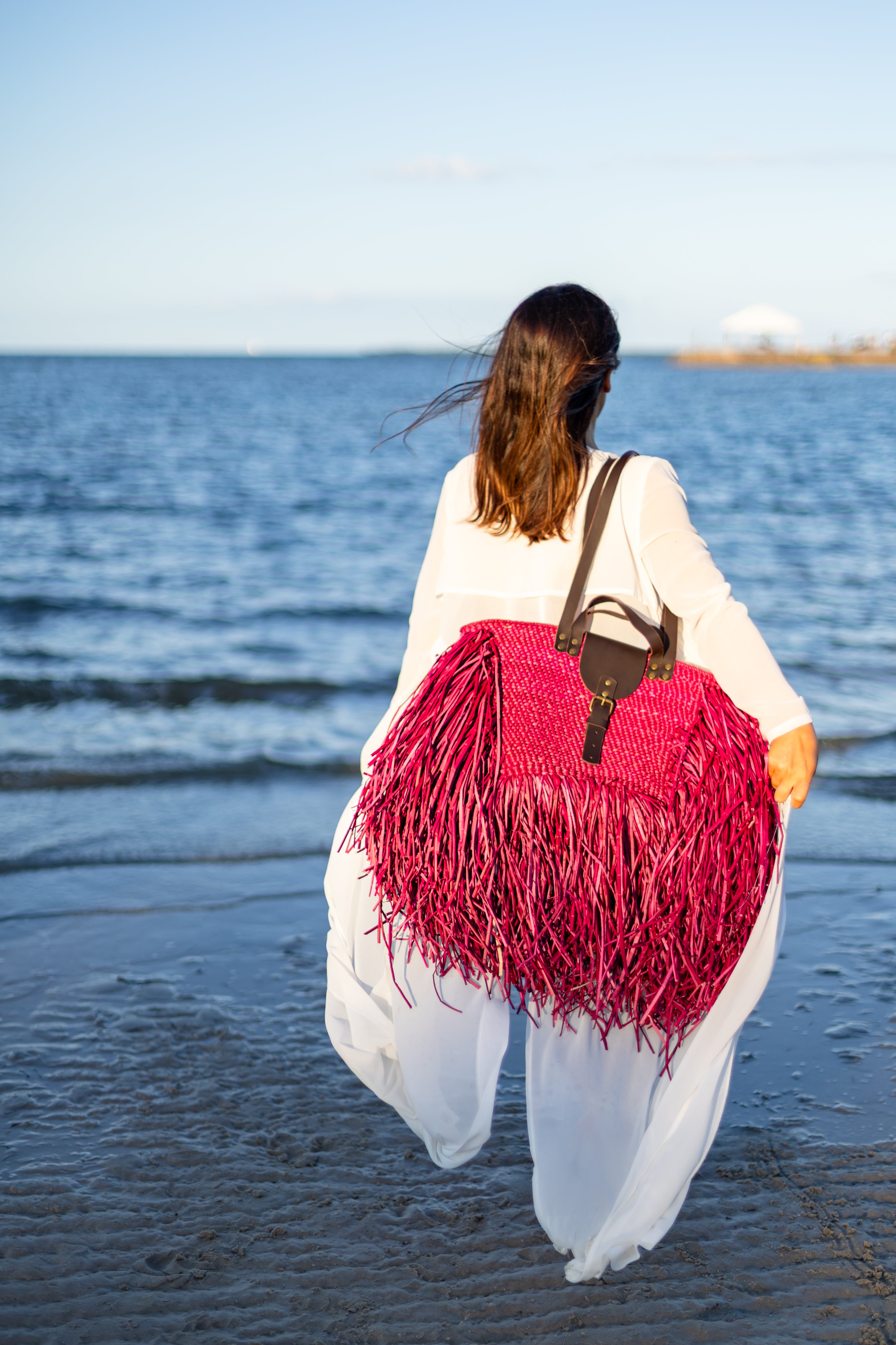 Woman walking on a beach with a pink and red fringed bag and white pants.