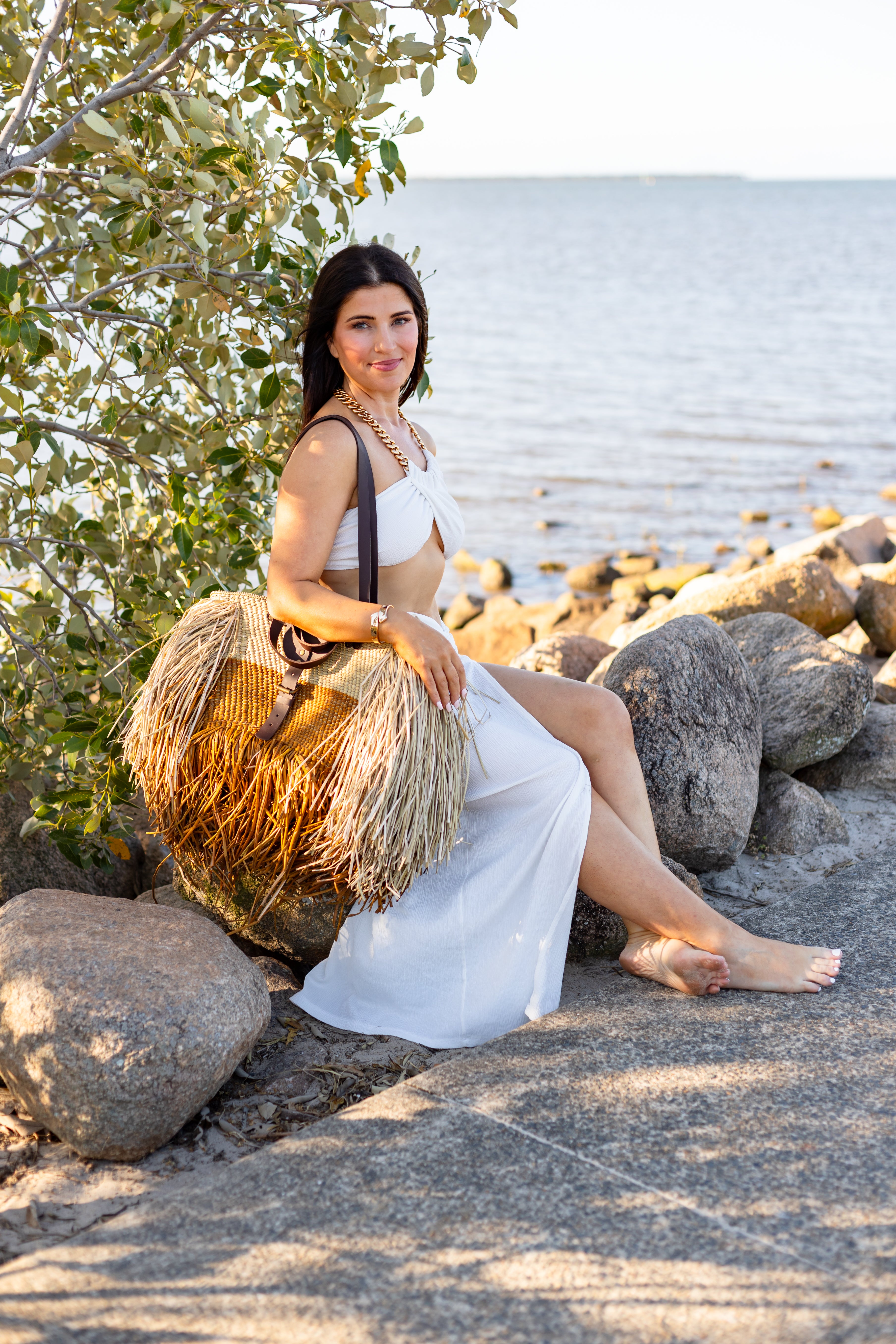 Woman in a white dress sitting on rocks by the water with fringed bag