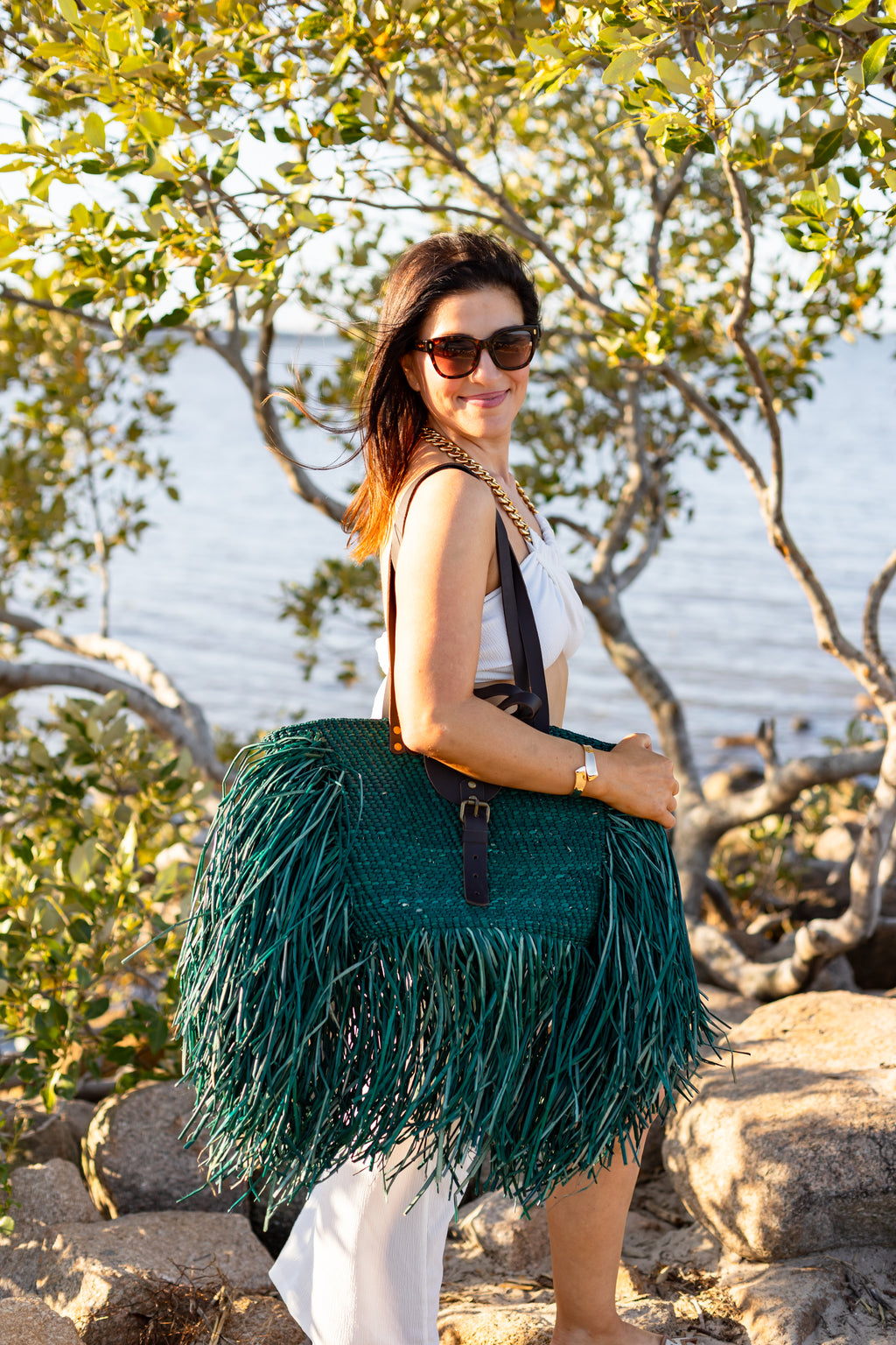 Woman holding a green fringed bag by a body of water with trees in the background