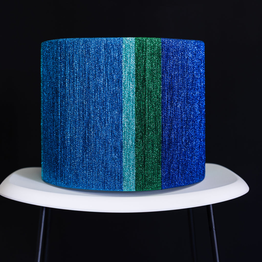Metallic blue and green thread wrapped drum lampshade on a white stand against a black background