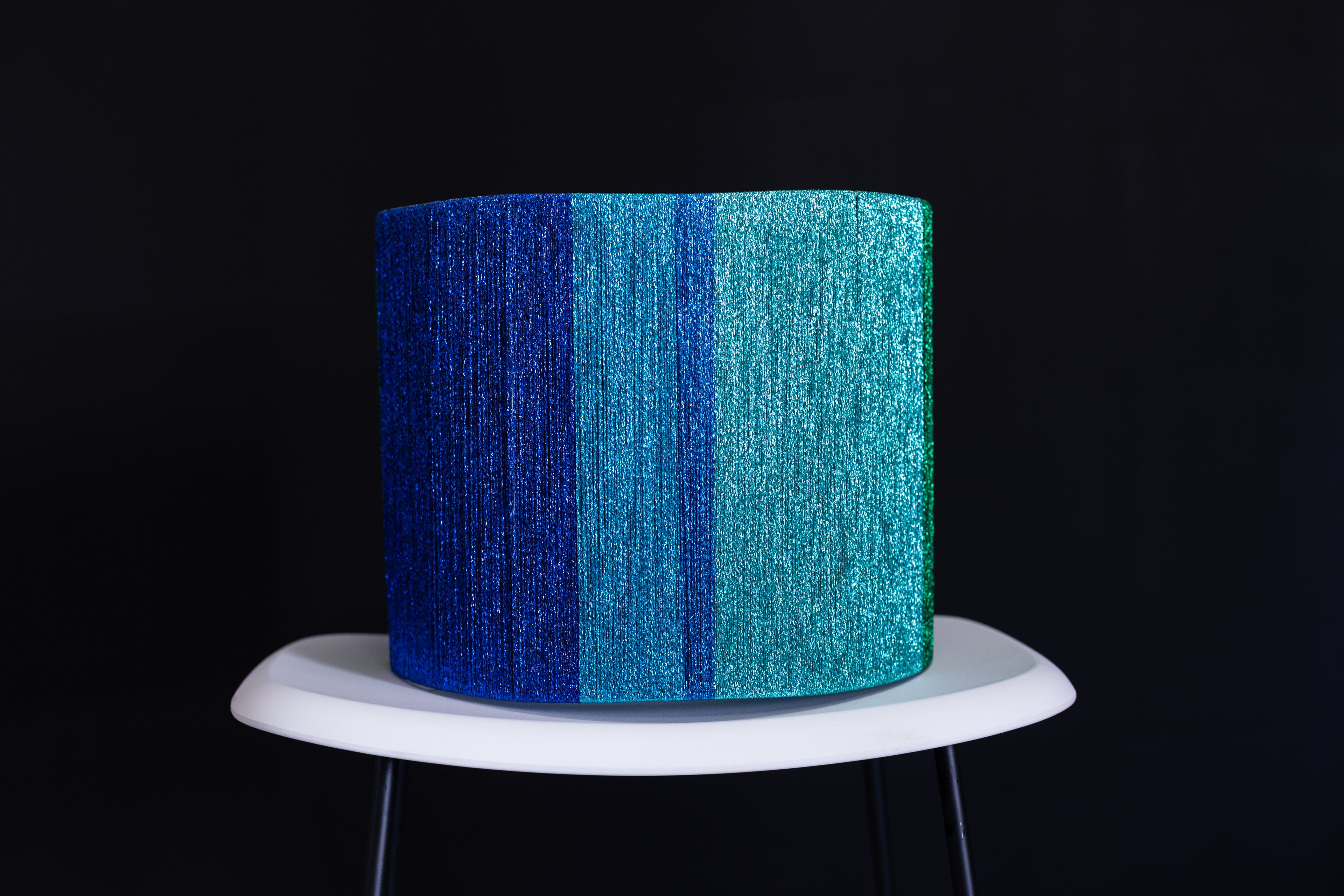 Metallic blue and green thread wrapped drum lampshade on a white stand against a black background