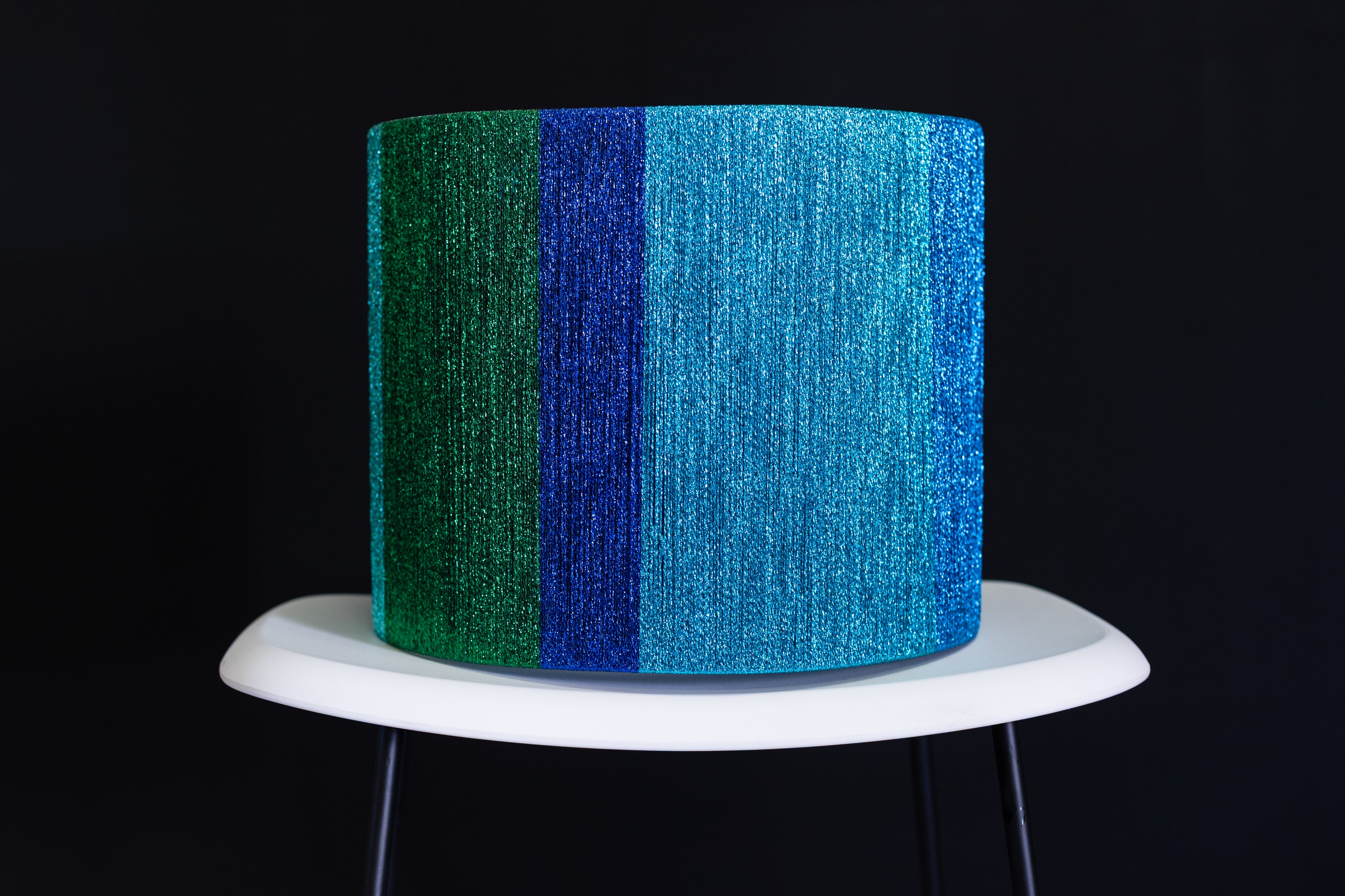 Metallic blue and green thread wrapped drum lampshade  on a white stand against a black background