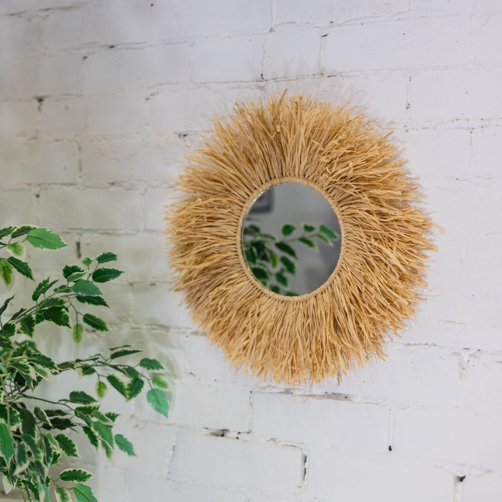 Round woven mirror on a white brick wall with green leaves.