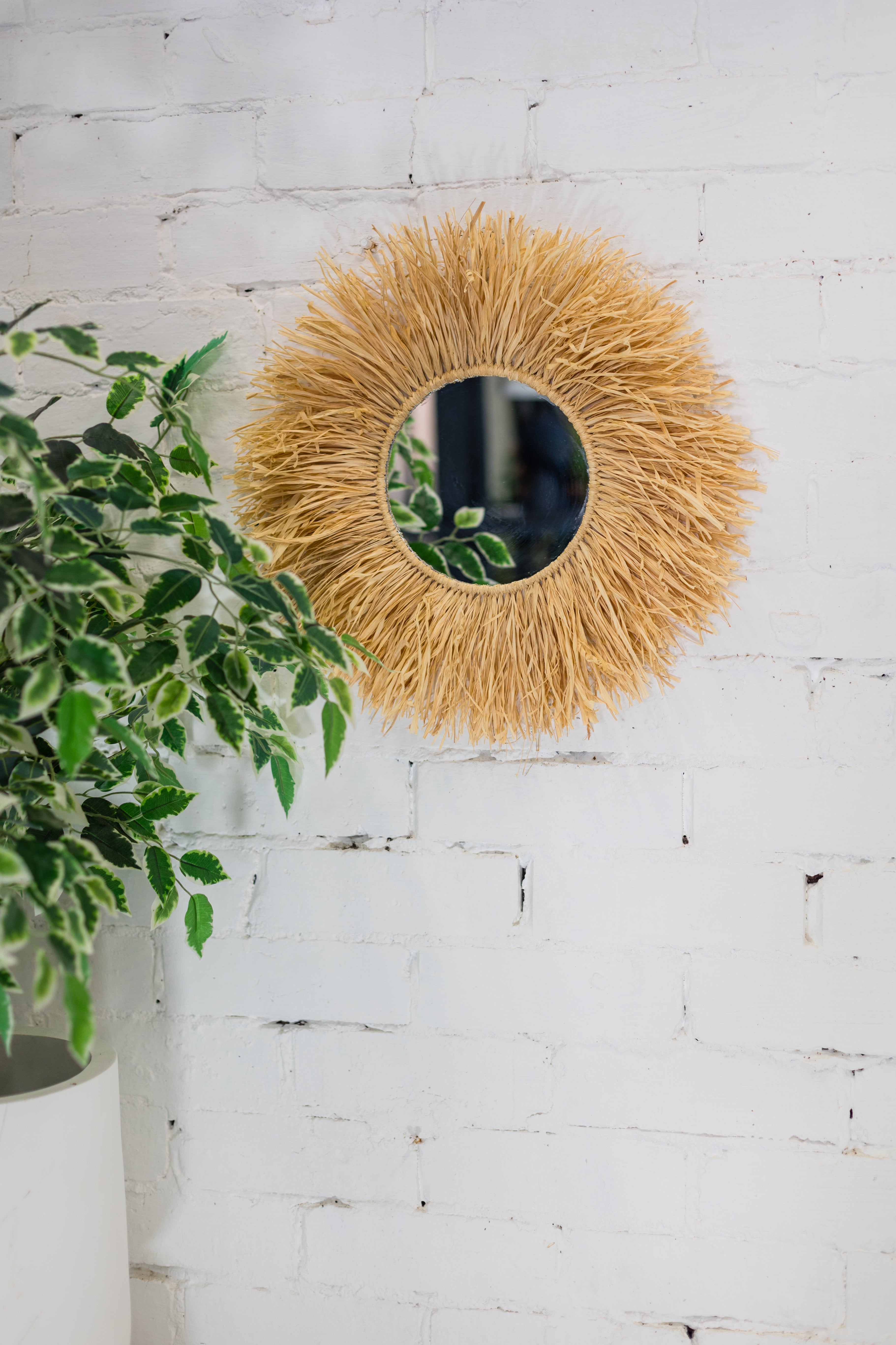 Round woven mirror on a white brick wall with a plant to the left.