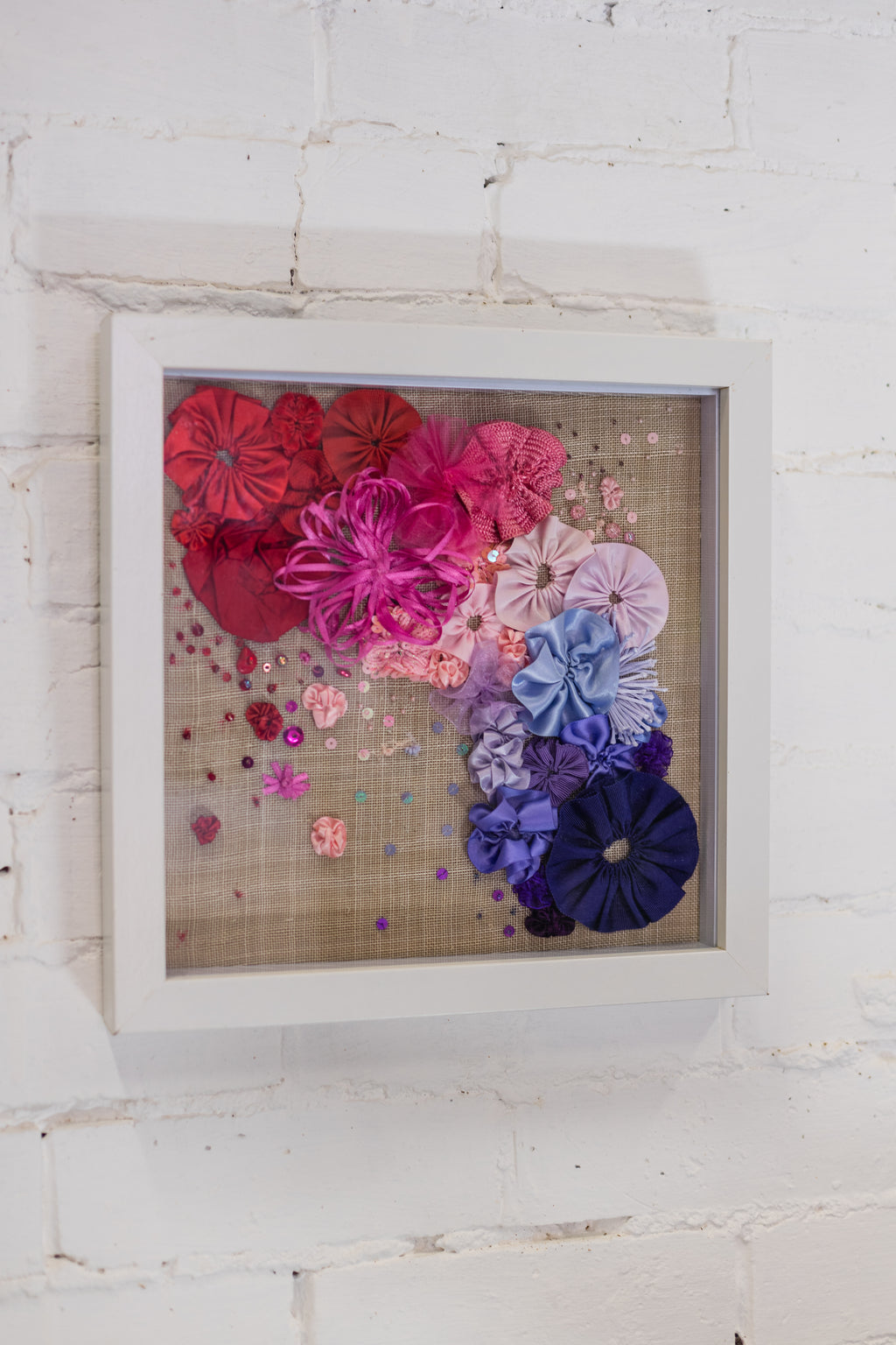 Decorative wall art with colourful ribbon flowers in a white frame on a white brick wall.