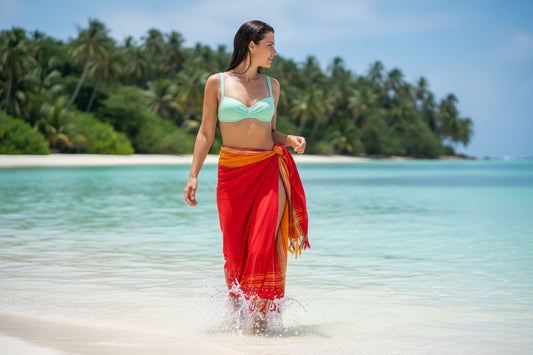 Woman in a green bikini top and red sarong walking on a tropical beach with clear blue water and palm trees in the background.